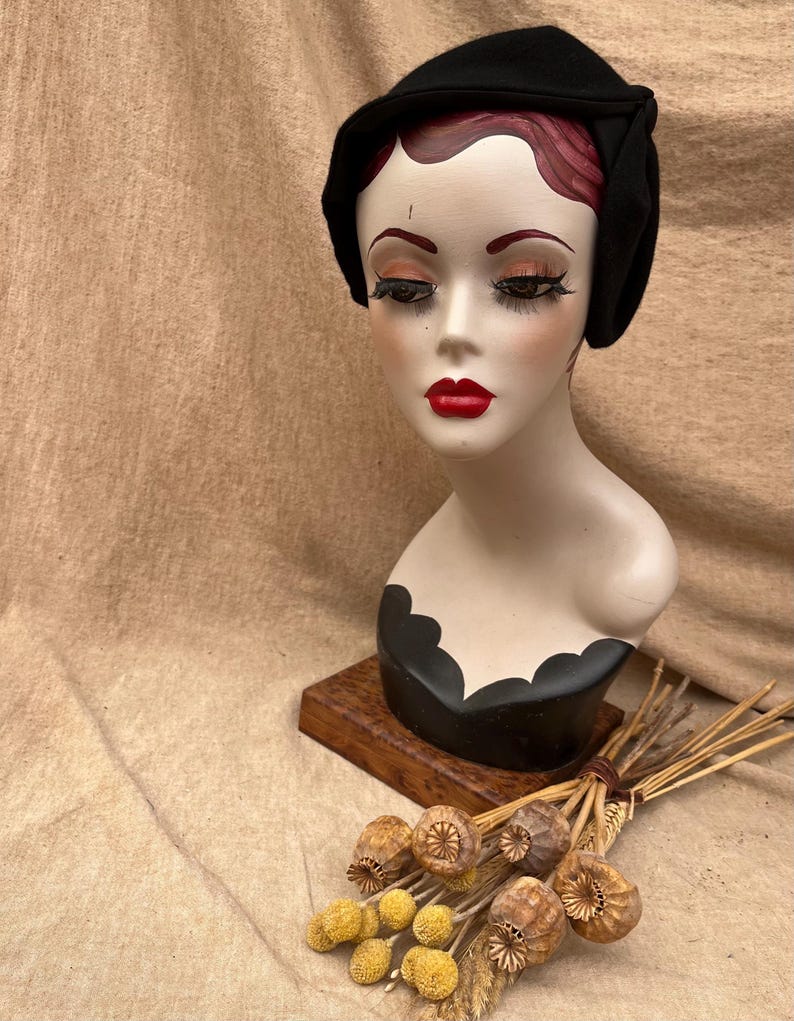 K&ouml;nnte beinhalten: Ein schwarzer Filzhut im Cloche-Stil mit schlichtem Design. Der Hut sitzt auf einem Mannequin-Kopf mit einer schwarz-wei&szlig; bemalten Basis.
