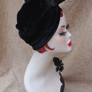 May include: A black velvet turban with a decorative embellishment. The turban is worn on a mannequin with red hair and a black lace choker.