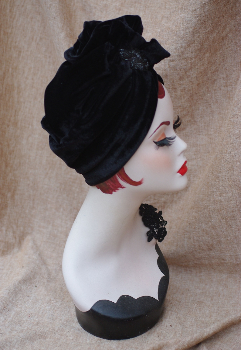 May include: A black velvet turban with a decorative embellishment. The turban is worn on a mannequin with red hair and a black lace choker.