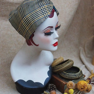May include: A mannequin head wearing a plaid turban style head wrap. The mannequin has red hair and red lipstick. The head wrap is brown, black, and white. The mannequin is on a black stand with a white base. The background is a beige fabric.