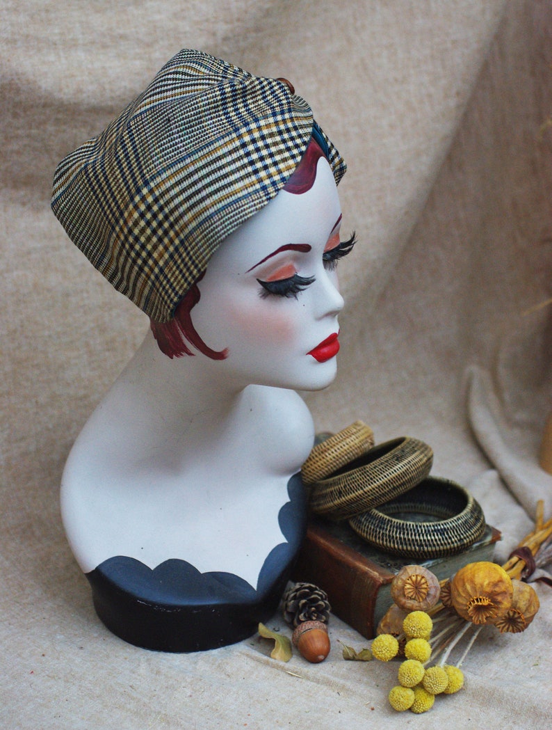 May include: A mannequin head wearing a plaid turban style head wrap. The mannequin has red hair and red lipstick. The head wrap is brown, black, and white. The mannequin is on a black stand with a white base. The background is a beige fabric.
