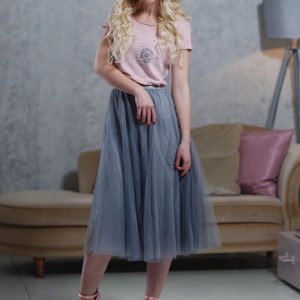 Peut inclure: Une femme portant un t-shirt rose avec une jupe en tulle grise et un accessoire pour cheveux fleuri. Elle se tient devant un canap&eacute; avec un coussin rose.