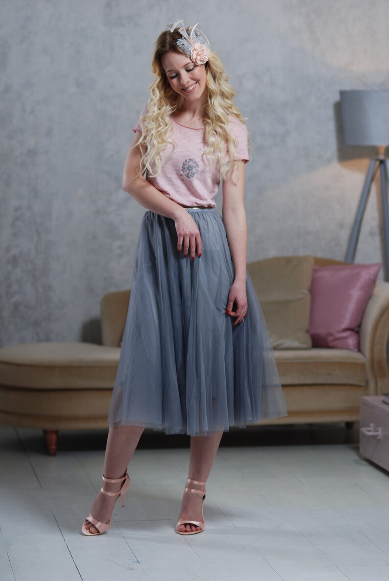 Peut inclure: Une femme portant un t-shirt rose avec une jupe en tulle grise et un accessoire pour cheveux fleuri. Elle se tient devant un canap&eacute; avec un coussin rose.
