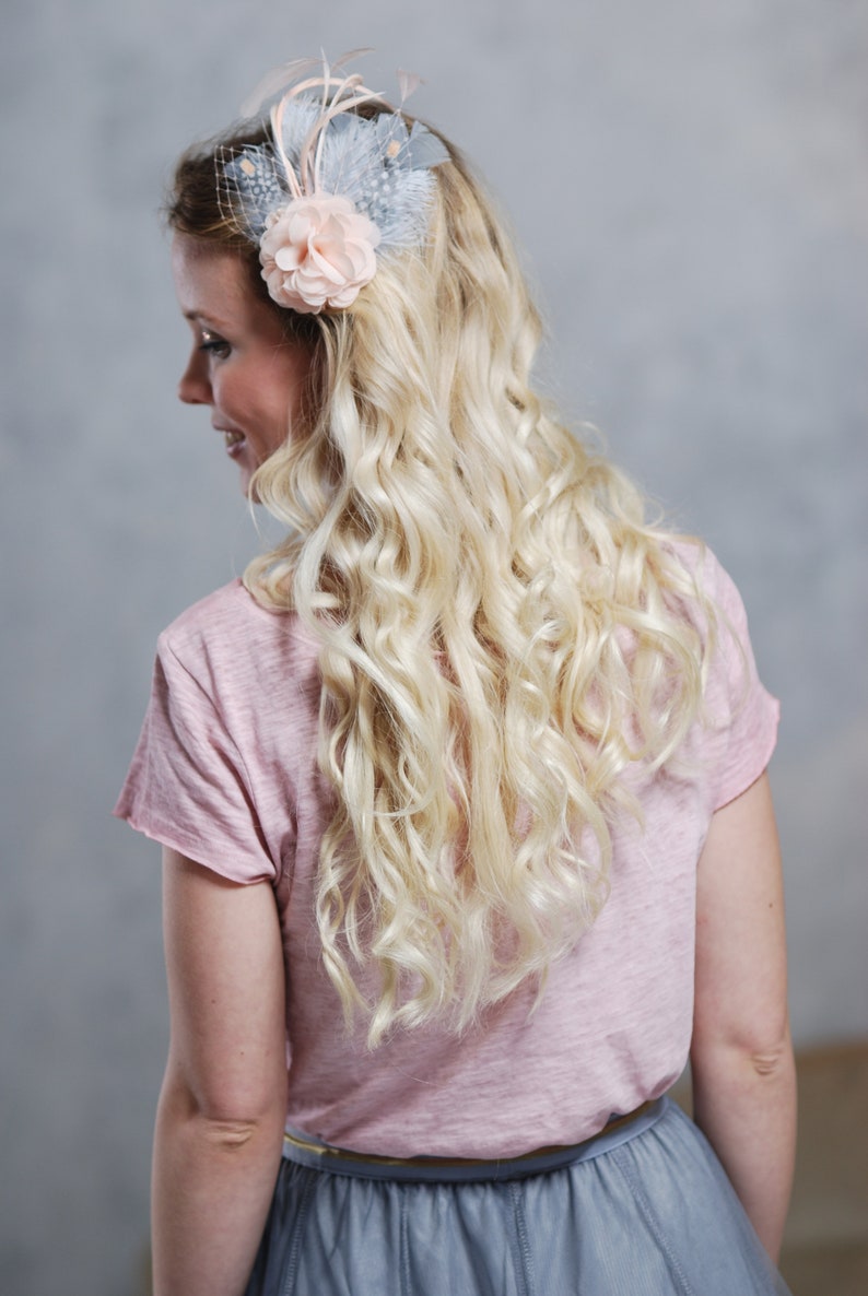Peut inclure: Une femme avec de longs cheveux blonds boucl&eacute;s portant un t-shirt rose et une jupe grise. Elle porte une barrette de cheveux en forme de fleur bleu clair et rose avec des plumes.