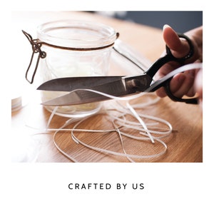 May include: Black metal scissors cutting a thin white ribbon, positioned in front of a clear glass jar with a metal latch closure. The text CRAFTED BY US is displayed below the image, which shows loose white ribbon strands on a wooden surface.