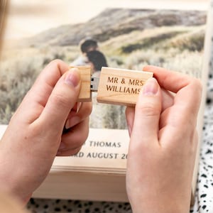 May include: A light-coloured wooden USB flash drive with the engraved text MR & MRS WILLIAMS. The drive is held in two parts, revealing the metal connector. In the background, a wooden box is visible with the text THOMAS and AUGUST 20.