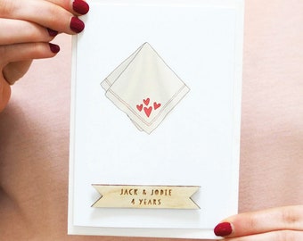 May include: A white greeting card featuring a central illustration of a folded white handkerchief with four small red hearts. Below the illustration, a wooden banner is attached with the engraved text "JACK & JODIE" and "4 YEARS".