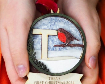 May include: A circular wooden ornament featuring a layered design with a red-breasted robin perched on a branch next to the letter T. The background shows a snowy scene, and a banner at the bottom reads THEA'S FIRST CHRISTMAS 2024.