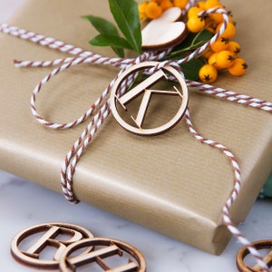 May include: A gold-wrapped gift box tied with brown and white baker's twine, featuring a wooden circular tag with the letter K, a wooden heart, and a sprig of green leaves with small yellow berries. Additional wooden letter tags lie nearby.