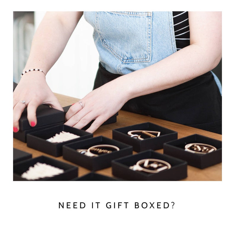 May include: Several small black square gift boxes arranged on a wooden surface, each containing a unique wooden ornament or jewelry piece. The text NEED IT GIFT BOXED? is displayed below the boxes, which feature various shapes including hearts and circles.