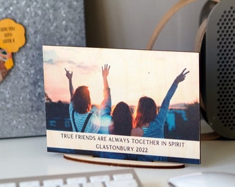 May include: A rectangular wooden plaque featuring a printed photograph of three people with their arms raised against a sunset sky. The text "TRUE FRIENDS ARE ALWAYS TOGETHER IN SPIRIT GLASTONBURY 2022" is printed in black at the bottom of the wooden board.