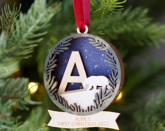 May include: A circular wooden Christmas ornament featuring a dark blue starry background, a large letter A, and a white bear silhouette. A banner at the bottom is engraved with the text ALBIE'S FIRST CHRISTMAS 2023. It hangs from a red ribbon.