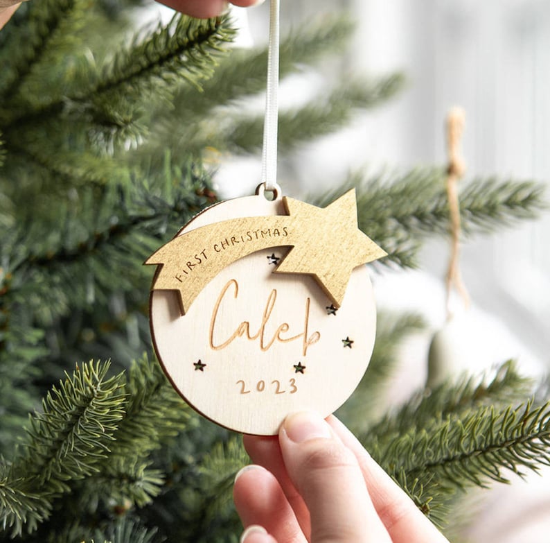 May include: A round wooden Christmas ornament featuring a gold-colored star and a banner with the text FIRST CHRISTMAS. The center is engraved with the name Caleb in script, the year 2023, and small star cutouts. It hangs from a white ribbon.