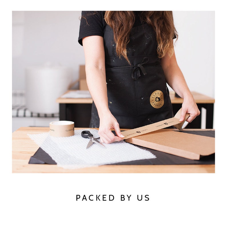 May include: A black apron with a circular gold logo, a roll of brown adhesive tape with black text, a pair of silver scissors, and a sheet of clear bubble wrap on a wooden table. A brown cardboard box is being sealed with the branded adhesive tape.