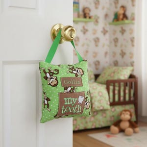 May include: A green fabric tooth fairy pillow featuring a repeating monkey pattern and white polka dots. The pillow has a green strap hanging from a brass door knob. It includes a brown rectangular patch with the name Collin and a pocket with the text my tooth.