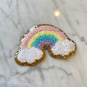 May include: A rainbow-colored felt patch with a gold glitter background and white fluffy clouds. The rainbow is made of pink, yellow, light blue, purple, and green felt.
