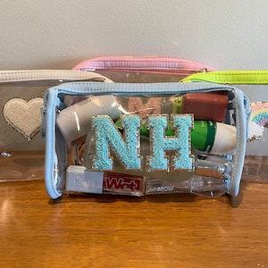 May include: A clear blue cosmetic bag with a zipper closure. The bag is filled with various beauty products, including a tube of sunscreen, a small bottle of hand sanitizer, and a tube of lip balm. The bag also features a large, blue and gold letter "NH" on the front.