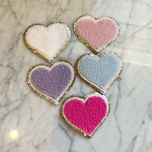 May include: Five heart-shaped patches with gold glitter trim. The hearts are made of a fluffy, fuzzy material and come in pink, white, light blue, purple, and hot pink.
