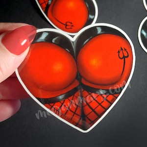 May include: A red heart-shaped sticker with a black and red design. The design features two red circles with a black outline and a black and red grid pattern. A black trident is on the right side of the sticker.