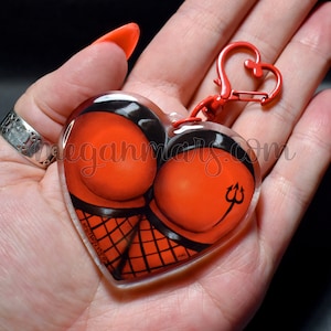 May include: A red heart-shaped keyring with a black and red illustration of a woman's backside with a devil's tail and trident. The keyring has a red metal clasp.