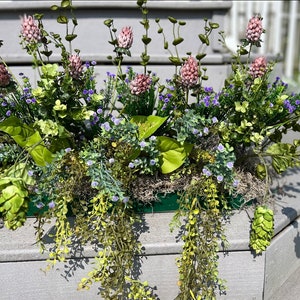 May include: A green planter filled with a variety of green plants and pink and purple flowers. The planter is sitting on a wooden surface.