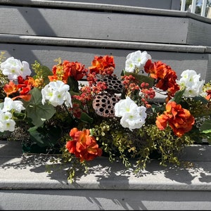 May include: A floral arrangement with white and orange flowers in a green planter. The arrangement is on a set of gray wooden steps.