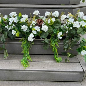 May include: A rectangular planter box overflowing with white petunia-like flowers, green ivy, and fern-like foliage. A dried seed pod adds a natural accent. The arrangement sits on gray wooden steps.