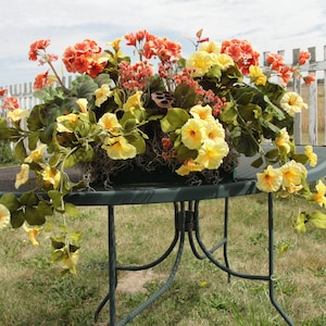 May include: A floral arrangement with yellow and orange flowers in a metal planter on a black metal table. The table is outdoors on a grassy lawn.