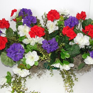 May include: A floral arrangement featuring red, white, and purple flowers. The flowers are arranged in a basket and are surrounded by green foliage.