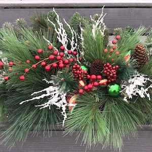 May include: A green pine garland with red berries, pine cones, and green and red ornaments. The garland is decorated with white frosted branches and is perfect for a Christmas centerpiece.
