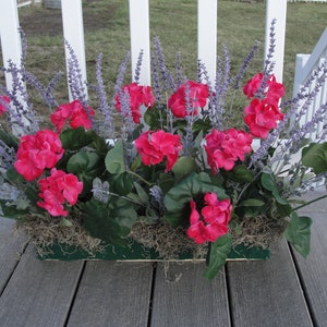 May include: A rectangular planter box filled with vibrant pink geraniums and purple lavender sprigs. The flowers are set in a bed of green moss and surrounded by green leaves. The planter is on a wooden deck.