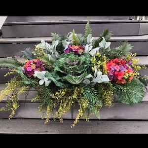 May include: A green cabbage plant with pink and orange flowers in a planter. The planter is filled with greenery and is sitting on a set of wooden stairs.