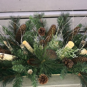 May include: A green planter filled with pine branches, pine cones, birch logs, and white twig sprigs. The planter is sitting on a wooden deck.