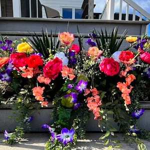 May include: A vibrant floral arrangement overflowing with colorful blooms. The display features large red peonies, orange orchids, yellow and pink roses, and trailing purple morning glories. Green foliage provides a lush backdrop for the bright flowers.
