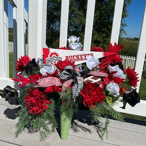 May include: A green and white floral arrangement with red, white, and black flowers. The arrangement includes a football and two pennants that say "Ohio State" and "Buckeyes".