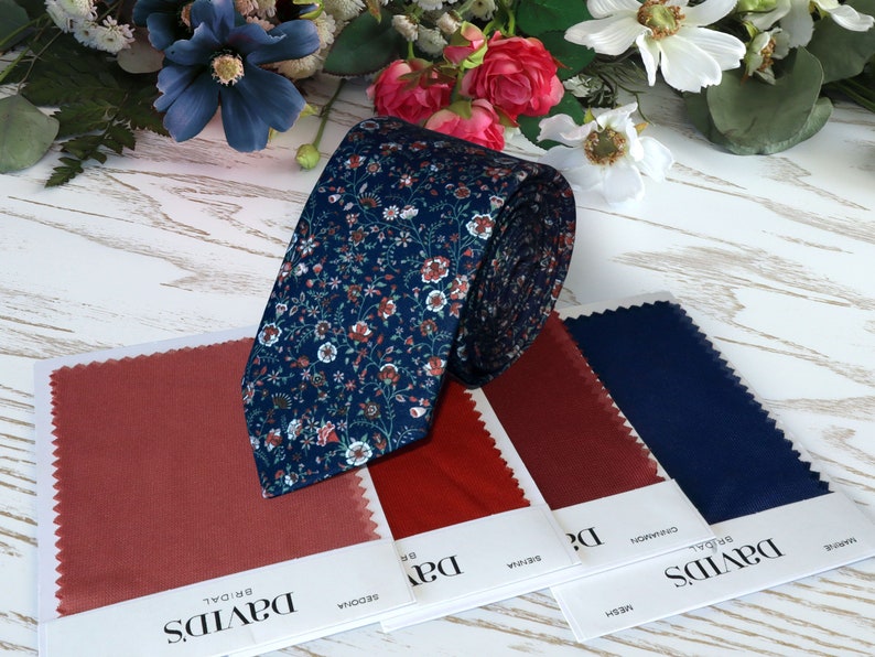 May include: A blue necktie with a floral pattern in shades of pink, white, and green. The tie is rolled up and displayed next to fabric swatches in shades of red and blue. The swatches are labeled with the brand name "David's Bridal" and the color names "Sedona", "Cinnamon", and "Navy".