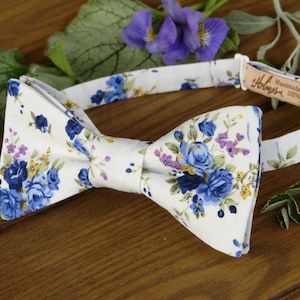 Puede incluir: Una pajarita blanca con un estampado floral azul y amarillo. La pajarita est&aacute; hecha a mano y es 100% algod&oacute;n.