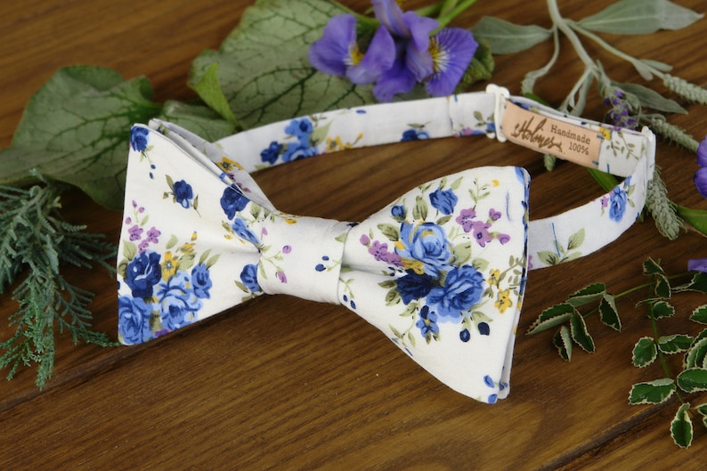 Puede incluir: Una pajarita blanca con un estampado floral azul y amarillo. La pajarita est&aacute; hecha a mano y es 100% algod&oacute;n.