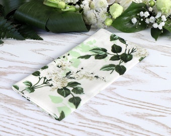 Può includere: Un tovagliolo di stoffa bianco con un motivo floreale verde. Il tovagliolo è piegato a metà e si trova su una superficie di legno bianco.
