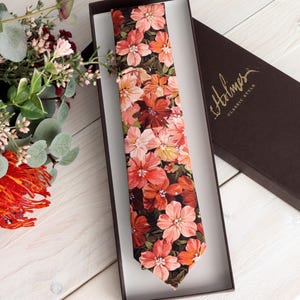 May include: A floral tie with a dark background and a pattern of red, orange, and pink flowers, presented in a brown box. The box has the brand name "Holmes" printed in gold. A bouquet of flowers is in the background.