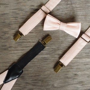 May include: A blush pink bow tie and suspenders set with brass-colored hardware. The suspenders have a black elastic back and are arranged on a weathered wooden surface. The bow tie is centered between the suspenders.