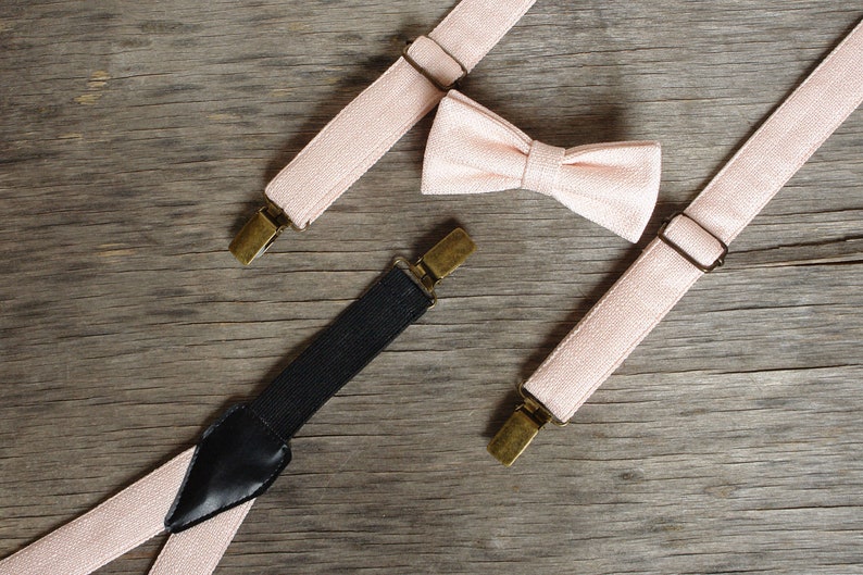 May include: A blush pink bow tie and suspenders set with brass-colored hardware. The suspenders have a black elastic back and are arranged on a weathered wooden surface. The bow tie is centered between the suspenders.