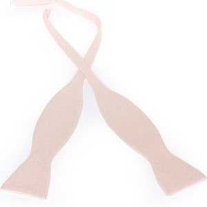 May include: A light beige bow tie, with a textured surface, is displayed against a white background. The bow tie is untied, with the two ends crossed over each other. The bow tie is a formal accessory.