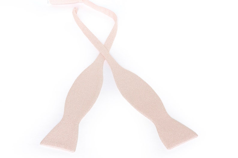 May include: A light beige bow tie, with a textured surface, is displayed against a white background. The bow tie is untied, with the two ends crossed over each other. The bow tie is a formal accessory.