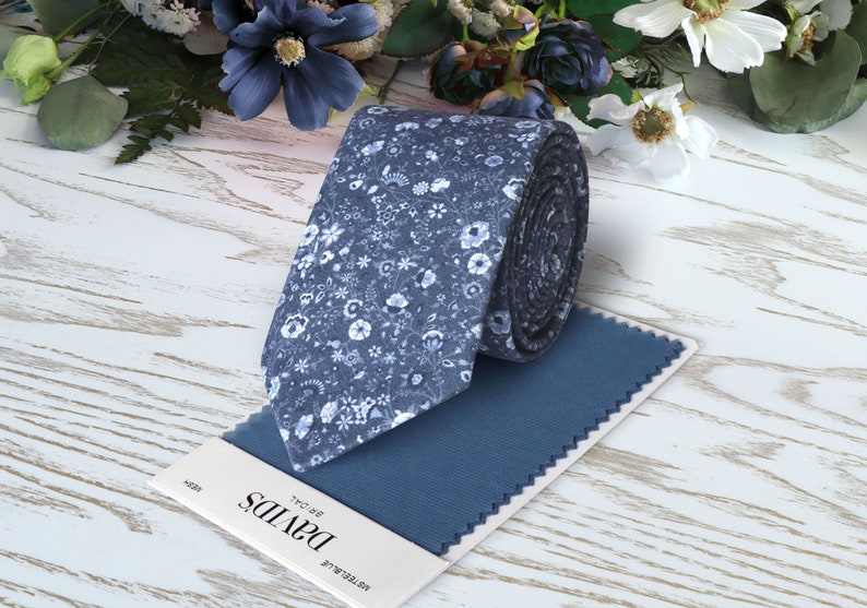 May include: A blue tie with a white floral pattern. The tie is rolled up and placed on a blue fabric swatch. The fabric swatch has the text "NOTRE-DAME" and "DAVIDS" printed on it.