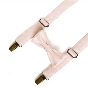 May include: A light pink bow tie and suspenders set. The bow tie is centered between the suspenders. The suspenders have metal clips and adjustable metal sliders. The set is made of a textured fabric.