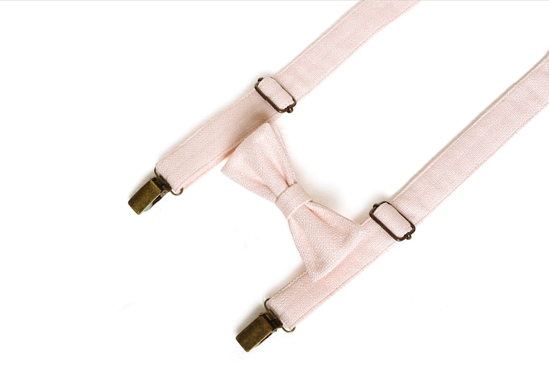 May include: A light pink bow tie and suspenders set. The bow tie is centered between the suspenders. The suspenders have metal clips and adjustable metal sliders. The set is made of a textured fabric.