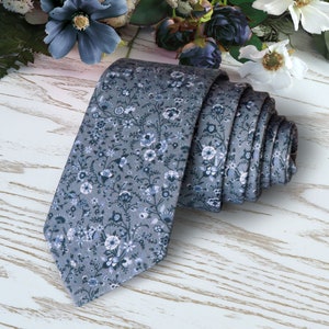 May include: A grey necktie with a white floral pattern. The tie is rolled up and lying on a white wooden surface.