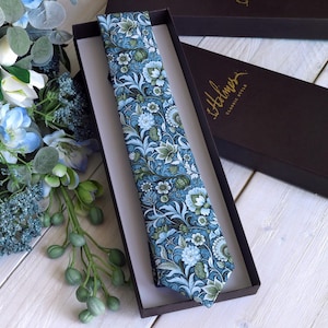 Könnte beinhalten: Eine Krawatte mit dichtem Blumenmuster in Blau-, Grün- und Weißtönen, präsentiert in einer dunkelbraunen rechteckigen Schachtel. Der Deckel liegt daneben und zeigt den goldenen Schriftzug Holmes Classic Style. Die Krawatte läuft spitz zu.