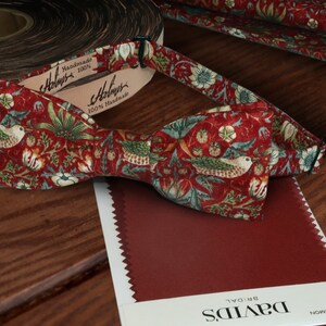 Peut inclure: Un nœud papillon rouge avec un motif floral représentant des oiseaux et des feuilles. Le nœud papillon est fait de tissu 100% fait main et est présenté sur une surface en bois avec un échantillon de tissu étiqueté "DAVID'S BRIDAL CRIMSON".